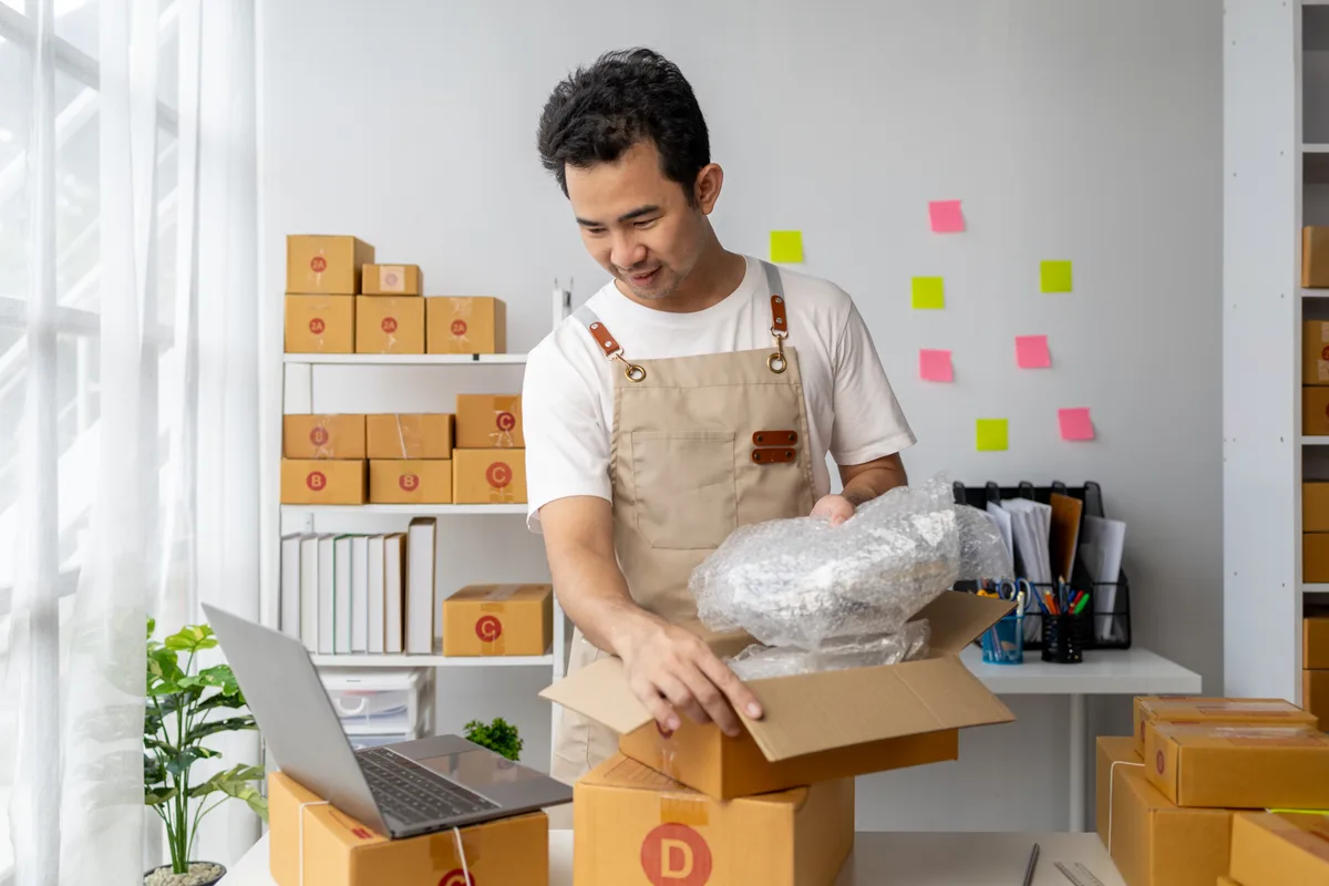 Online seller packing an order at a home workstation with shipping labels and a laptop showing a sales dashboard